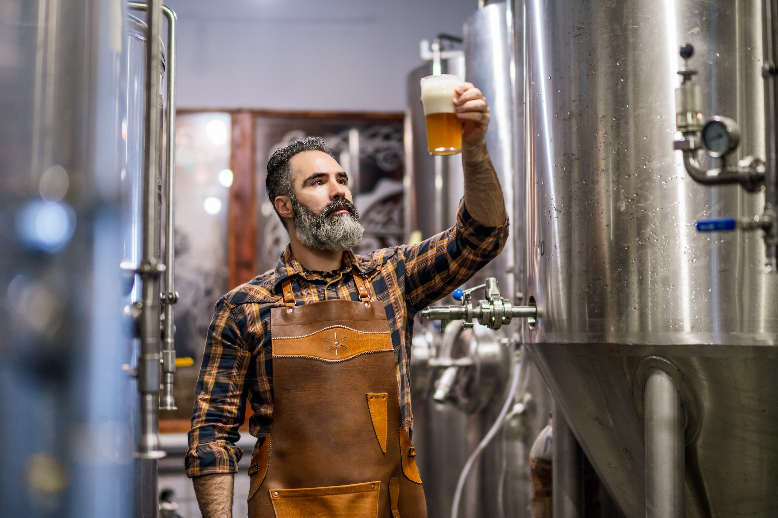 man in a brewery production facility inspecting a glass of frothy pasteurized beer in a clear glass